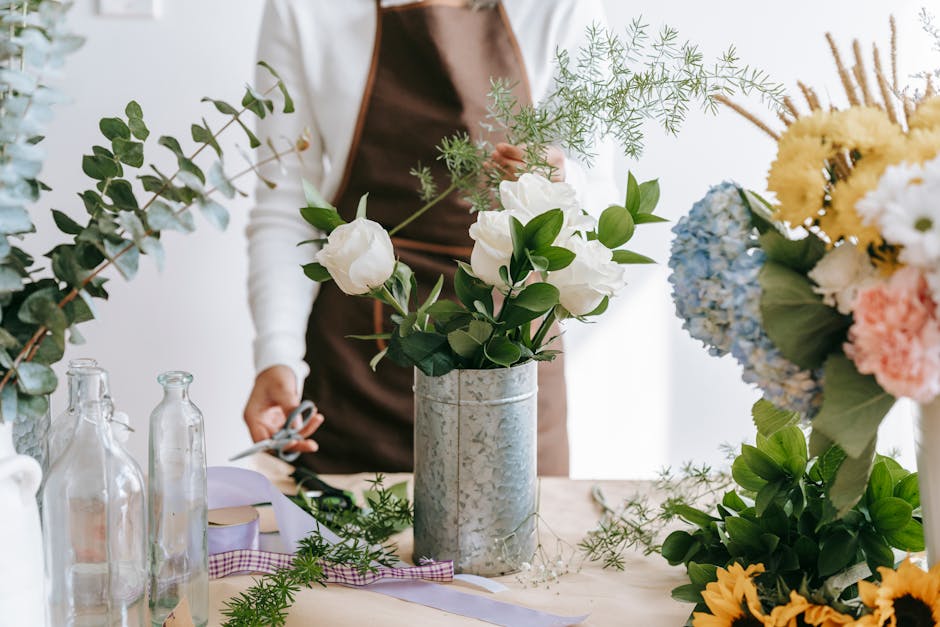 A person wearing a brown apron and white long-sleeved shirt arranging white roses and green foliage in a tall silver metal vase on a wooden table. Surrounding the vase are various flower arrangements,