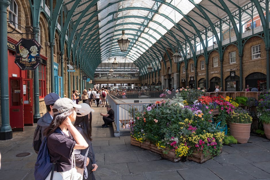 An indoor market with a high, arched glass ceiling supported by green metal framework, illuminated by hanging lanterns. In the foreground, three people wearing hats and dark clothing are standing and