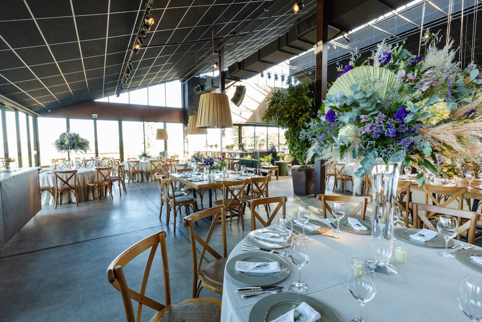 A spacious, modern event venue with large floor-to-ceiling windows allowing natural light to illuminate the room. In the foreground, a tall, clear glass vase holds a floral arrangement featuring laven