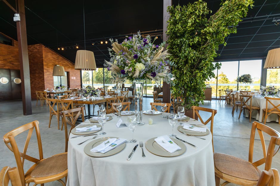 A large round table set for a formal event in an elegant event space, featuring a tall glass vase centerpiece filled with an arrangement of fresh flowers including white and purple blooms, complemente