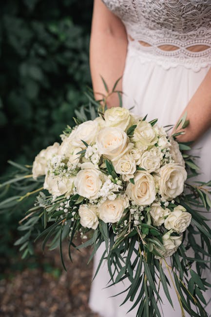 A close-up of a bride holding a lush floral bouquet featuring creamy white roses, complemented by smaller white blossoms and sprigs of greenery with elongated, dark green leaves. The bouquet is arrang