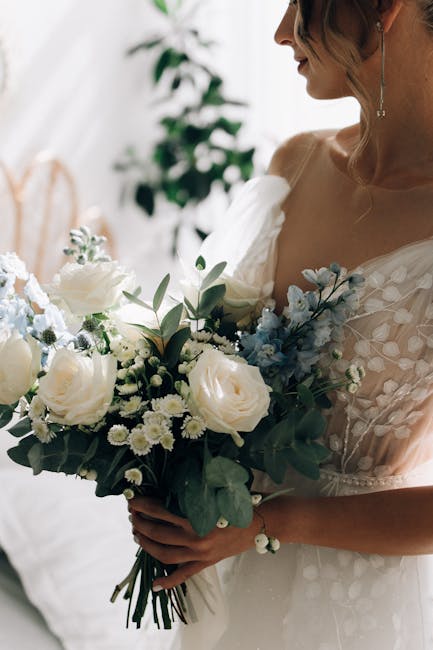 A woman dressed in a white wedding gown with delicate floral embroidery holds a large, fresh flower bouquet featuring white roses, small white daisies, and light blue flowers complemented by green fol