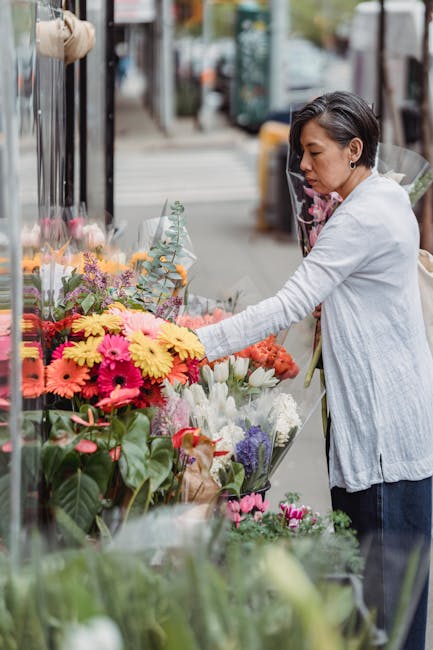 A woman selecting fresh flowers from an outdoor floral display on a sidewalk, featuring a vibrant arrangement of pink, yellow, and purple blooms including gerbera daisies and lavender, with green foli