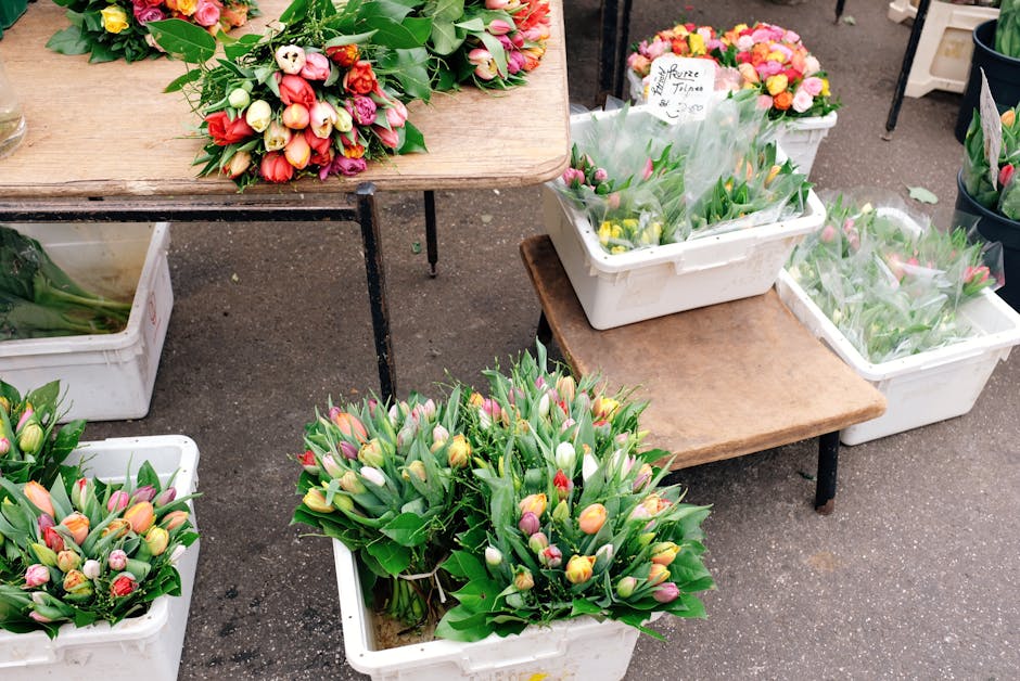 A flower stall displaying fresh tulip bouquets with a variety of pastel pink, cream, yellow, and purple tulips, arranged in white plastic containers and placed on wooden tables with black metal suppor