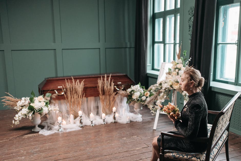 A woman seated on an antique-style upholstered chair in a softly lit room with green paneled walls and large windows, holding a floral arrangement of peach roses and light pink blooms. In front of her
