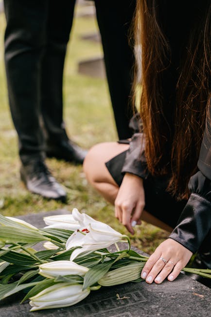 A person kneeling on the ground, arranging a floral tribute with white lilies and green foliage at a memorial site. The lilies, featuring large petals and prominent stamen, are wrapped in simple green