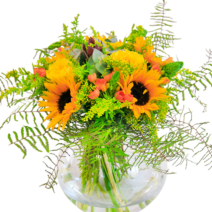 This Putney floral arrangement instantly dazzles with an air of golden optimism-sunflowers at its core, their bright petals arching around dark, velvety centers to conjure the light-filled promise of summer along the Thames. The sunny theme is echoed by plush, buttery yellow garden roses, each bloom swirling open with an inviting softness that brings a gentle fragrance reminiscent of Putney markets in full bloom. Glossy red hypericum berries nestle between the flowers like little jewels, their vibrant tones enlivening the subtle greens and elevating the design with lighthearted charm. The foliage is lush and dynamic: feathery fern fronds cascade naturally, evoking a sense of wild beauty found in Putney Heath or along Lower Richmond Road, while airy filler flowers and light eucalyptus leaves add freshness and balance. The clear, round glass vase lets every stem shine, filtering sunlight through green stalks and pure water, emphasizing the bouquet's pure, local vitality. Up close, the mix of textures-soft petals, crisp greens, smooth berries-invites touch and admiration. Intended as a tribute to happiness, this bouquet suits birthday celebrations, milestone anniversaries, or brightening a Putney home with the familiar warmth and charm only flowers crafted in SW15 can provide.