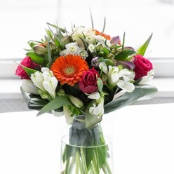 A visual testament to balance and artistry, this stunning bouquet brings together the vibrant spirit of Putney's community and nature's grace. The arrangement's core features creamy and fiery orange gerberas, their soft, almost halo-like petals embracing vivid centers that call to mind lantern-lit evenings by the Thames. Deep fuchsia roses add luxurious depth and a whisper of romance, while spirals of white alstroemeria and tiny spray roses create a gentle brightness, echoing the soft beauty found in Putney's beloved parks. Wisps of pale pink and petite white blooms emerge from tangles of foliage, weaving layers of color and texture that evoke abundance and generosity. The foundation is a cradling of paddle-shaped aspidistra leaves, broad and glossy, grounding the bouquet in lush tranquility much like leafy walks along Lower Richmond Road. Smaller, pointed green leaves interlace and add dynamic structure. The entire composition stands elegantly in a tall glass vase, where sturdy stems lend a sense of natural honesty and freshness. Diffused light streams in, highlighting the nuanced palette and giving the flowers an almost ethereal glow-perfect for a Putney brunch, a gallery opening, or welcoming a friend into a new space. Each aspect of this arrangement reflects the thoughtful creativity of Flowers Putney's design, infusing every bloom with optimism and care.